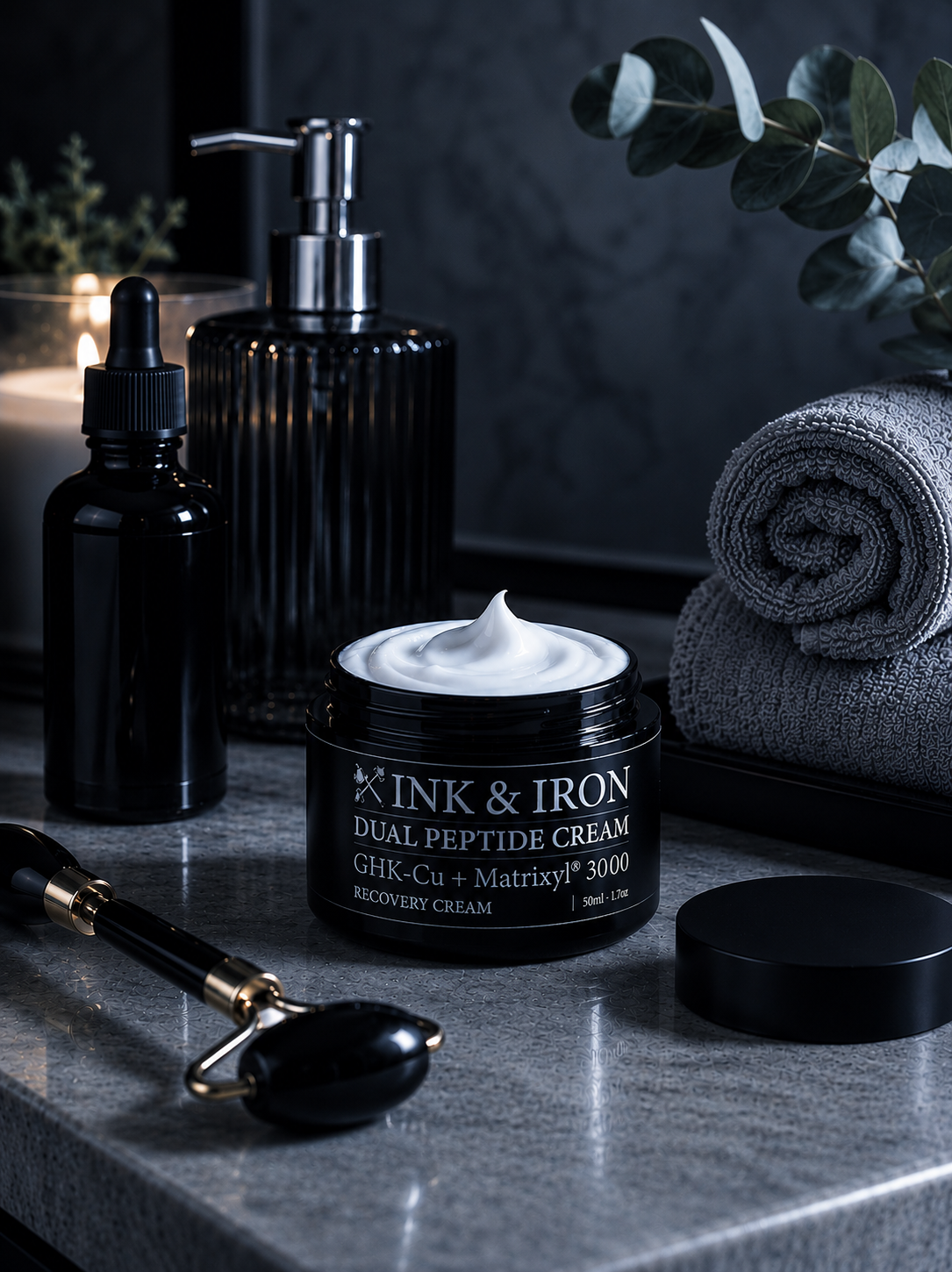 A jar of INK & IRON Dual Peptide Cream sits on a gray stone surface with its lid beside it. The scene includes a black pump bottle, a small black dropper bottle, a black jade roller, rolled gray towels, and greenery in a dark, spa-like setting.