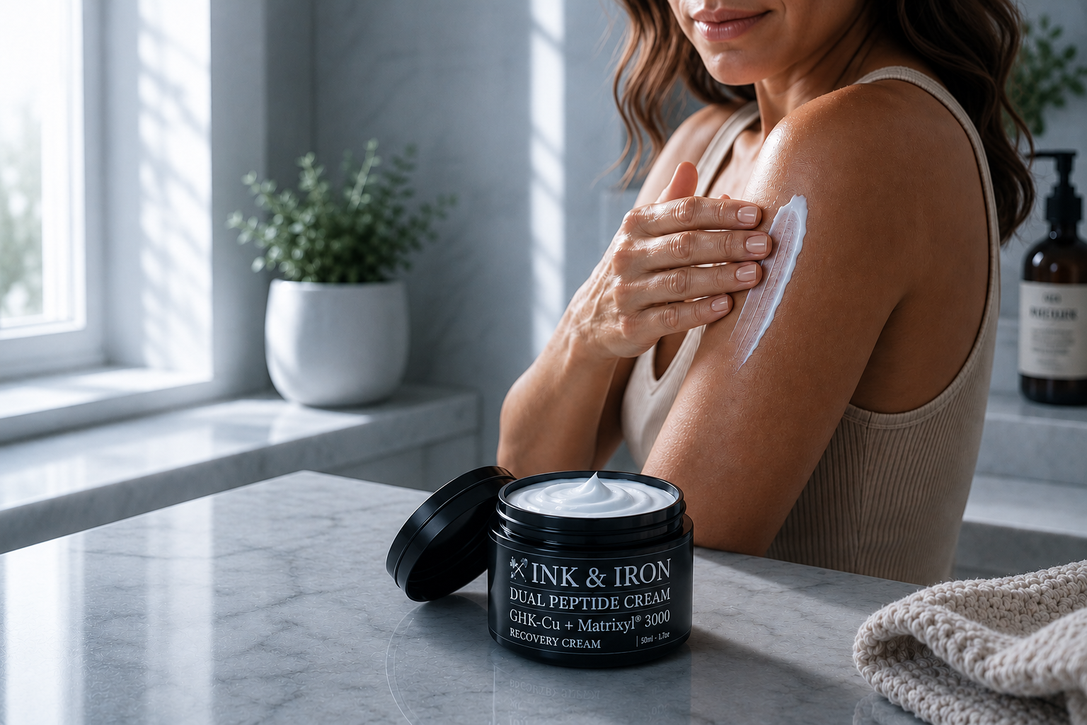 A woman applies a white body cream from an open black jar labeled INK & IRON RECOVERY CREAM to her upper arm in a bright bathroom setting.