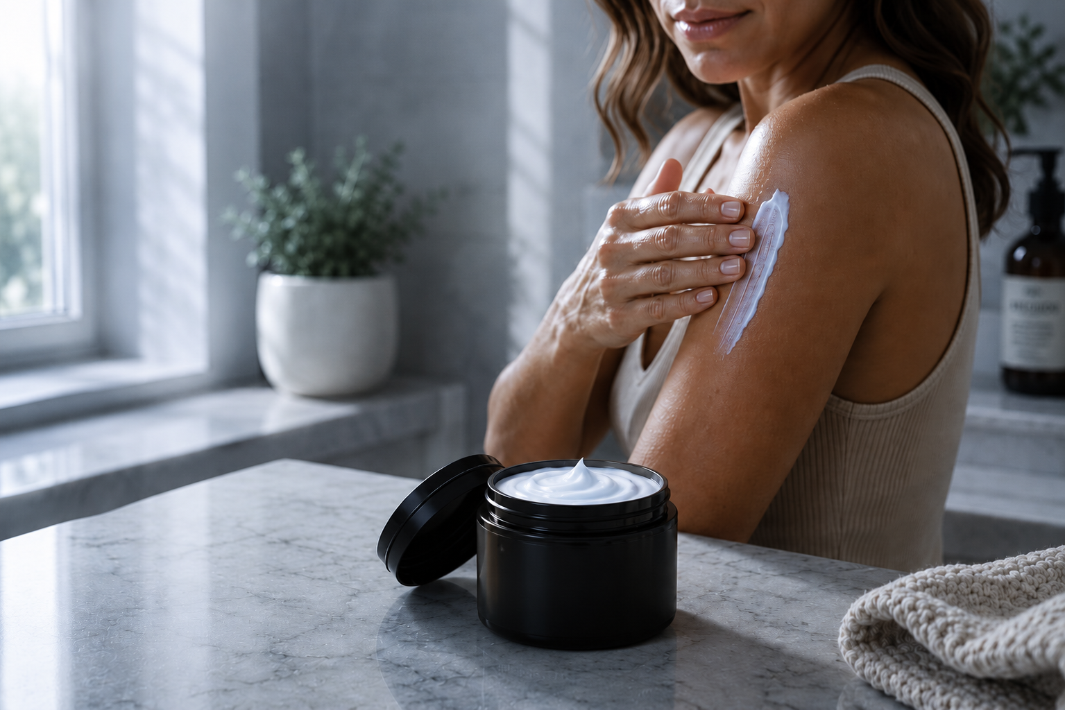 A woman applies white body cream from an open black jar onto her upper arm in a bright bathroom setting.